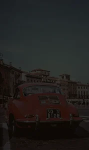 A photo of a red car with old buildings in the background, captured in Verona, Italy. The image has a CSS filter with 20% brightness, making it significantly less bright, almost black, compared to the original.