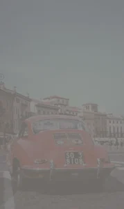 A photo of a red car with old buildings in the background, captured in Verona, Italy. The image has a CSS filter with 20% contrast, giving it a softer, more grayish look compared to the original.
